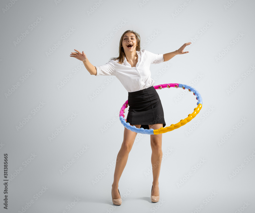 Like in childhood. Woman in office clothes training with hoop on grey studio background. Businesswoman training in motion, action. Unusual look for sport, new activity. Sport, healthy lifestyle. Like in childhood. Woman in office clothes training with hoop on grey studio background. Businesswoman training in motion, action. Unusual look for sport, new activity. Sport, healthy lifestyle.