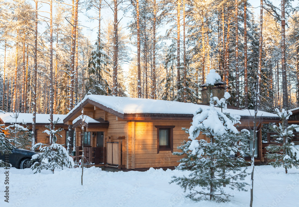 One-storey house made of wooden beams among the trees of the winter forest. Treetops lit by low sun