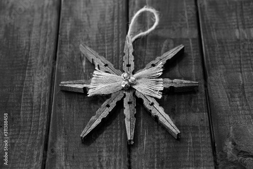 Black and white image. Homemade wooden star with a plaid bow and bells. Placed on top of dark wood floor.