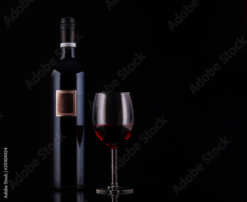 Closed red wine bottle with empty label and glass of wine on black background with reflections. Concept sales, discount price. Photo for advertising