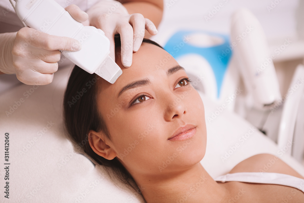 Attractive young Asian woman having her skin cleansed by cosmetologist using ultrasound cavitation apparatus