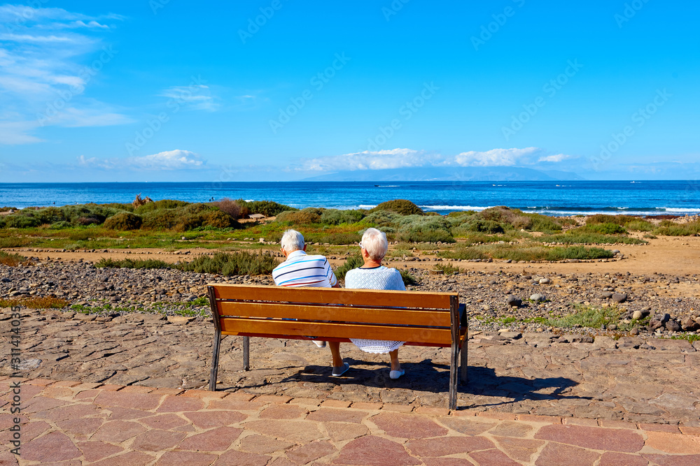 Elderly couple of tourists enjoy a rest on a bench with an beautiful ocean view on a summer sunny day. Tourism. Resort on Canary islands. 
