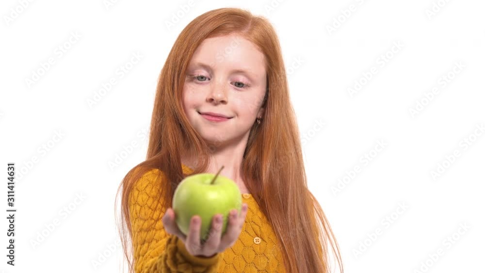 Close-up of a beautiful red haired young model posing at camera. Charming girl is smiling and offering juicy fresh green apple. Isolated, on white background Close-up of a beautiful red haired young model posing at camera. Charming girl is smiling and offering juicy fresh green apple. Isolated, on white background