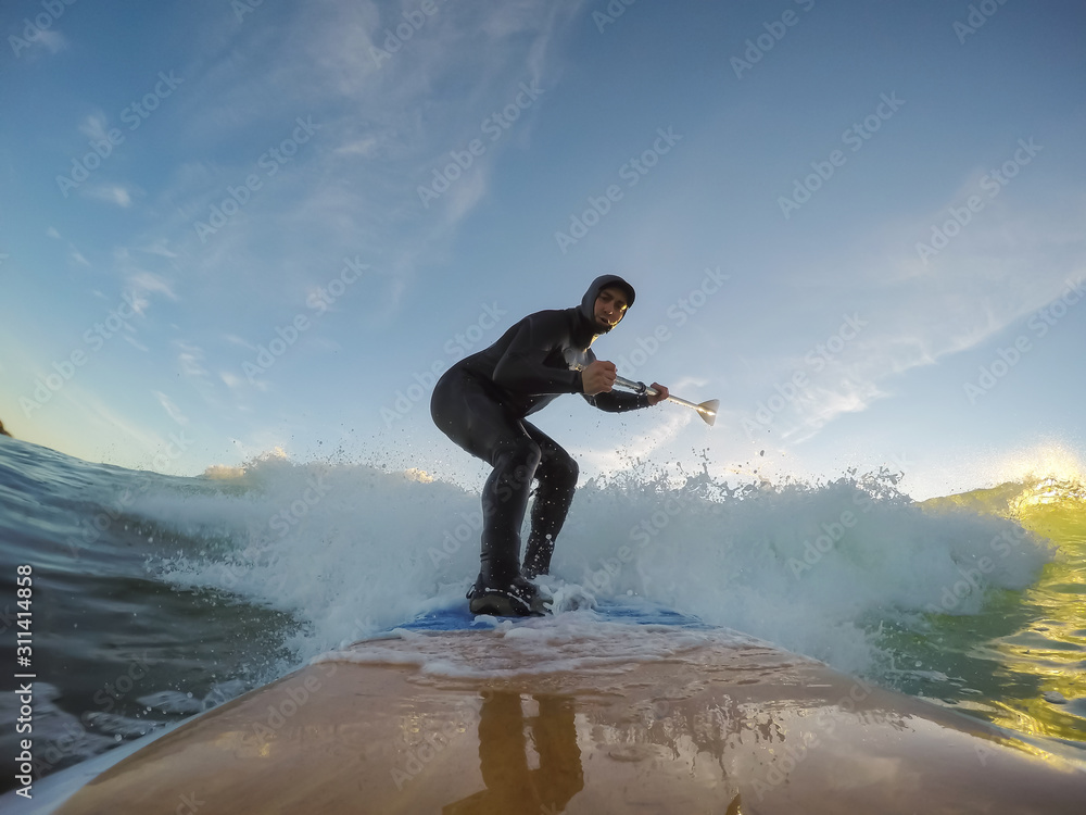 Obraz premium Adventurous Man Surfer on a paddle board is surfing in the ocean during a sunny sunset in Fall Season. Taken in Long Beach, Tofino, Vancouver Island, BC, Canada.