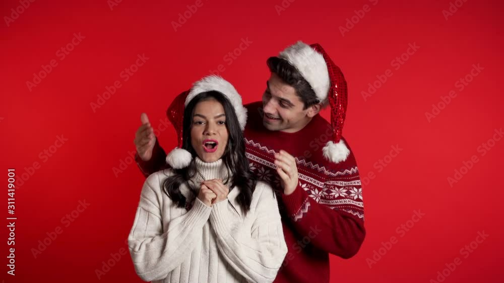 Man closes eyes of his beloved girlfriend before surprise her. Couple in Santa hat on red studio background. Love, holiday, happiness concept. Man closes eyes of his beloved girlfriend before surprise her. Couple in Santa hat on red studio background. Love, holiday, happiness concept.