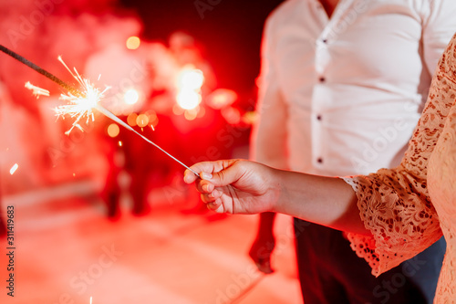 A crowd of young happy people with sparklers in their hands during celebration. Red sparklers at the wedding - bride, groom and guests holding lights in hand. Sparkling lights of bengal fires. Smoke.