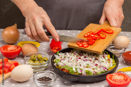 Sliced vegetables in pan. Hands put tomatoes in pan. Olives, eggs, peppers, tomatoes and onions on the table..