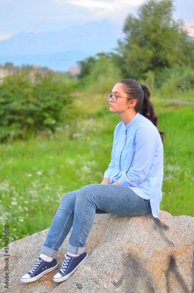 A young girl wearing glasses in a blue shirt and jeans sits on a large stone in a green clearing. Selective focus. A young girl wearing glasses in a blue shirt and jeans sits on a large stone in a green clearing. Selective focus.
