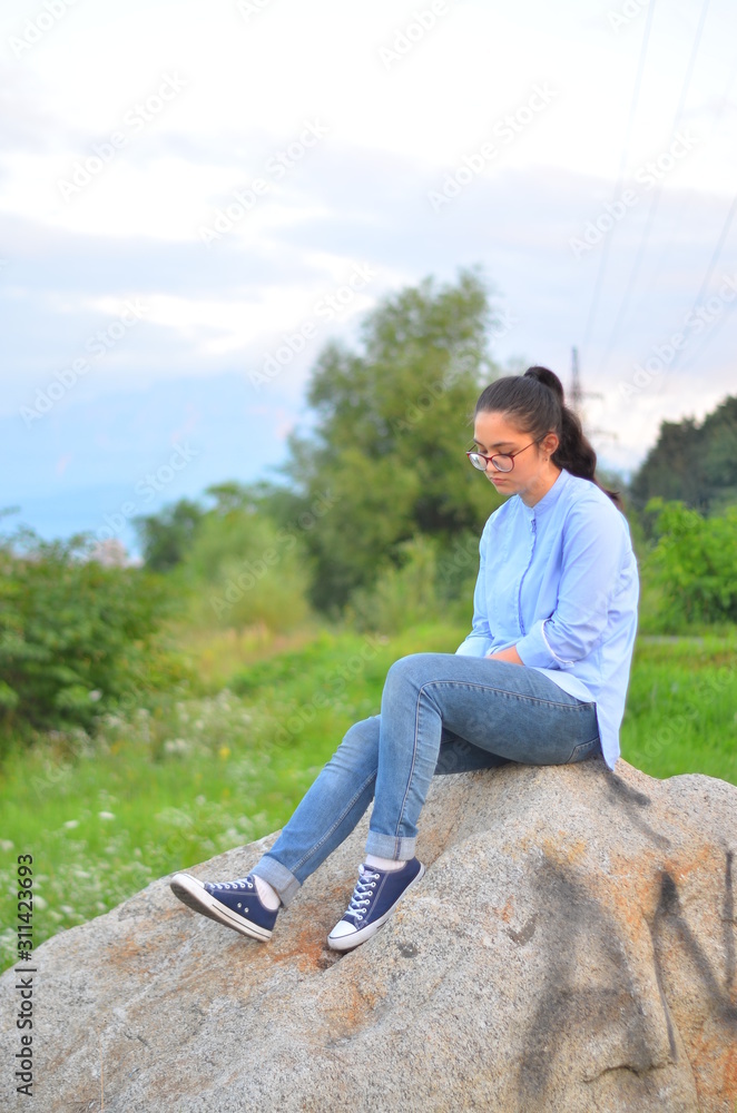 A young girl wearing glasses in a blue shirt and jeans sits on a large stone in a green clearing. Selective focus.