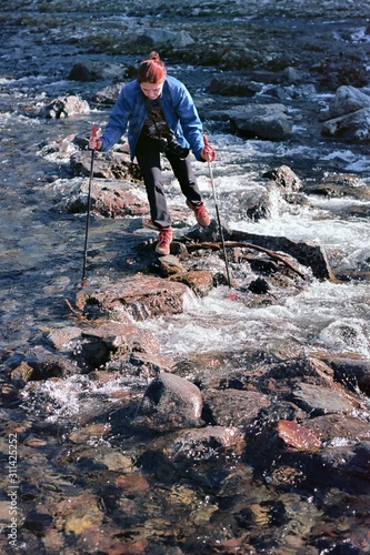 girl traveler crossing a mountain fast river over a crossing over rocks, Nordic walking sticks, Hiking