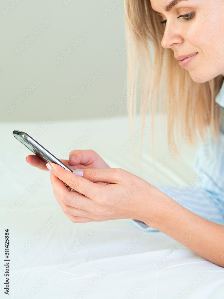 Joyful blonde European girl laying on the bed and looks at the phone screen. Close Up vertical photo Joyful blonde European girl laying on the bed and looks at the phone screen. Close Up vertical photo