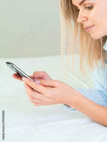 Joyful blonde European girl laying on the bed and looks at the phone screen. Close Up vertical photo