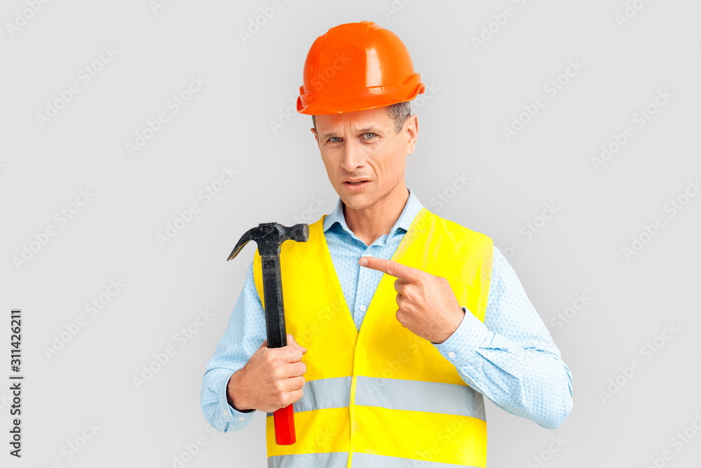 Construction. Mature man in hardhat and uniform standing isolated on white pointing at hammer concerned