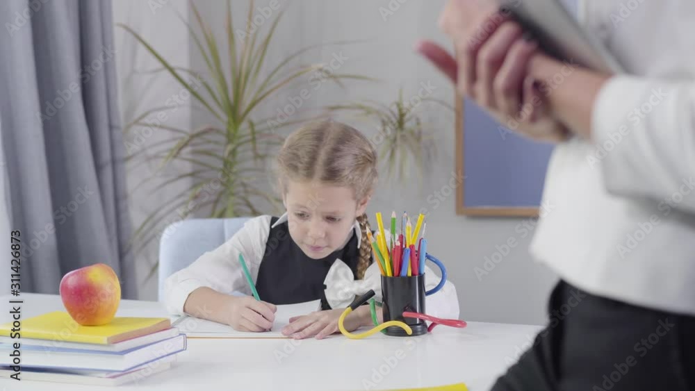 Charming smart Caucasian schoolgirl making i got an idea gesture and writing down in exercise book. Unrecognizable woman standing at the foreground on the right. Focused on child. Charming smart Caucasian schoolgirl making i got an idea gesture and writing down in exercise book. Unrecognizable woman standing at the foreground on the right. Focused on child.