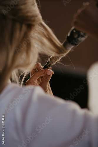 Bride twists the hair with a curling iron at home. Preparation of the bride. Close up. Women doing hairstyle herself and using straightener on beautiful long curl hair. Beauty and haircare concept