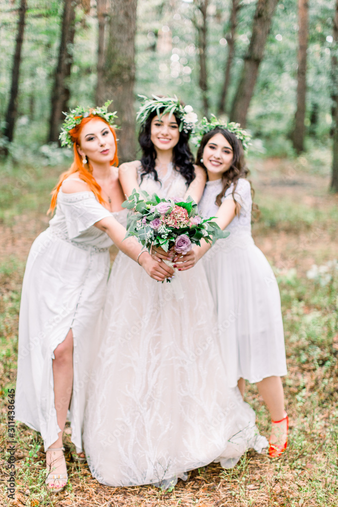 Beautiful bride in stylish rustic dress and wreath is holding flower bouquet in her hands. Two pretty bridesmaids in white dresses and wreaths are hugging the bride. Rustic wedding in the forest