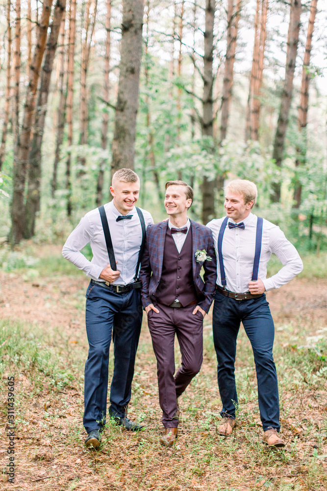 Cheerful stylish groom and groomsmen stand side by side, posing in the forest. Wedding shoot outdoors Cheerful stylish groom and groomsmen stand side by side, posing in the forest. Wedding shoot outdoors