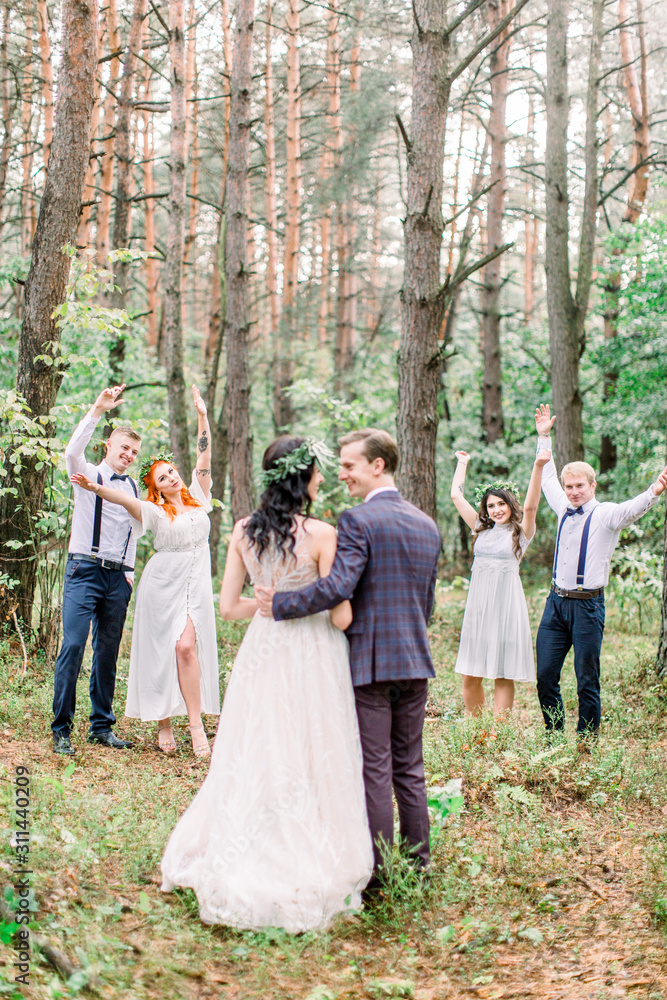 Fototapeta premium Stylish rustic wedding couple with bridesmaids and groomsmen at forest on wedding day. Back view of groom and bride, embracing and kissing. Their friends smiling and holding their hands on the air.