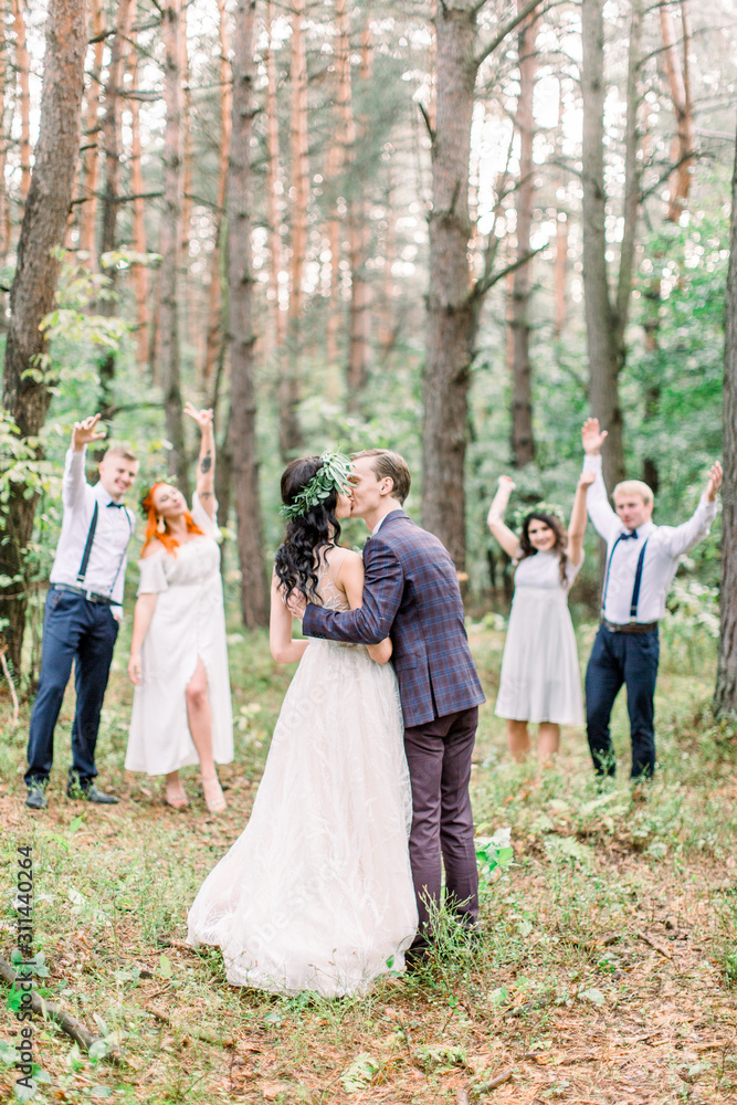 Stylish rustic wedding couple with bridesmaids and groomsmen at forest on wedding day. Back view of groom and bride, embracing and kissing. Their friends smiling and holding their hands on the air.