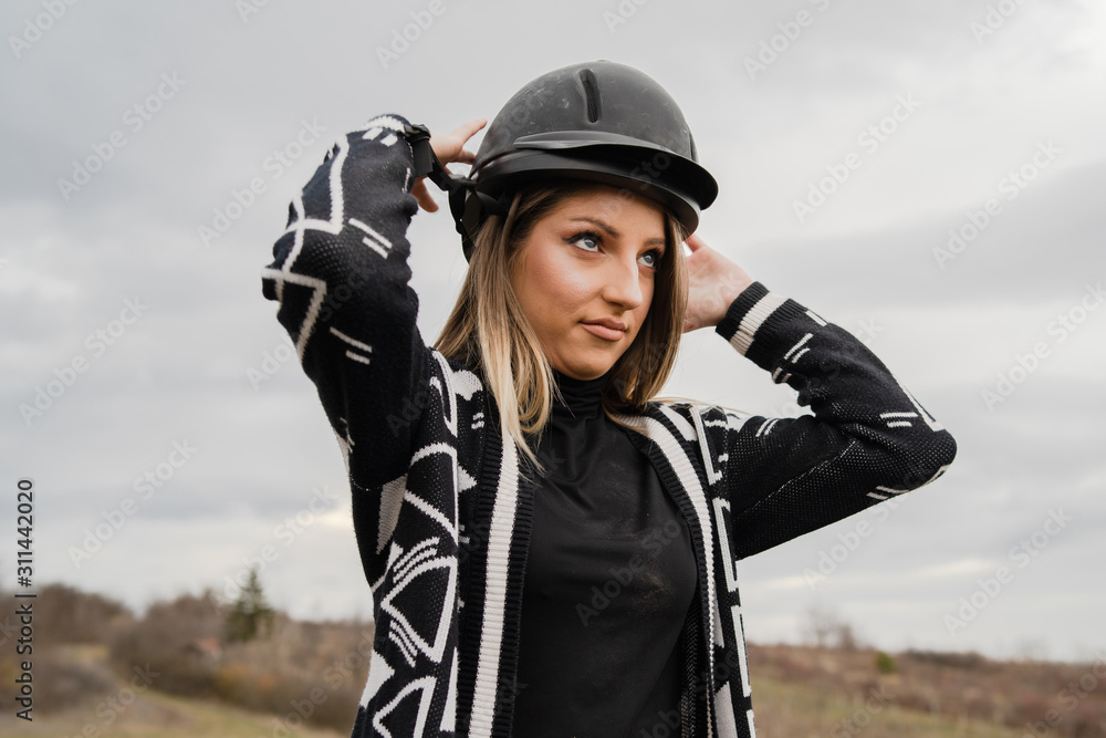 Young beautiful blonde caucasian woman female adjusting black protective helmet against a cloudy gray sky in winter or autumn day wearing sweater