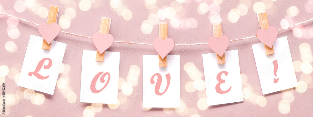 LOVE - Clothes pegs with wooden hearts and paper notes hang on rope isolated on soft pastel pink texture with bokeh lights background banner panorama long, with space for text