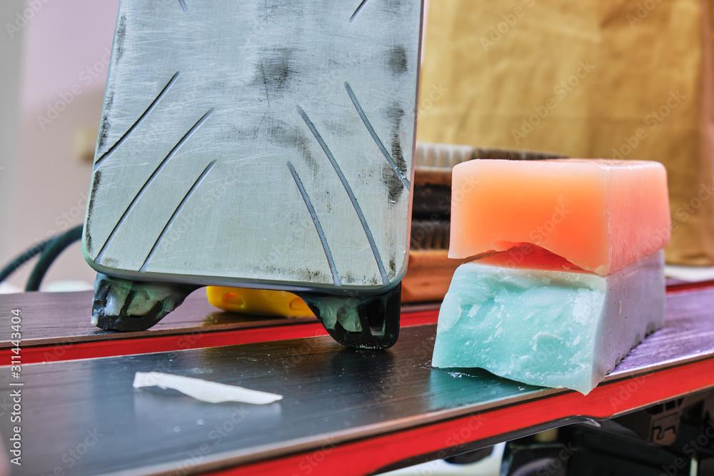 Ski wax iron and wax bars, colored orange and teal for different temperatures, laid on top of a pair of skis. Ski base tuning concept.