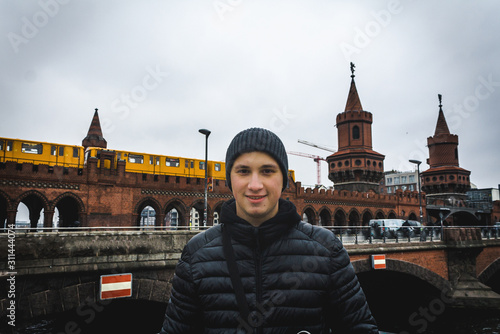 Young man smilling with Oberbaum Bridge and yellow train on background in cloudy weather in Berlin. Travel and tourism in Berlin concept. Europe travel in winter. Student and youth travel concept.