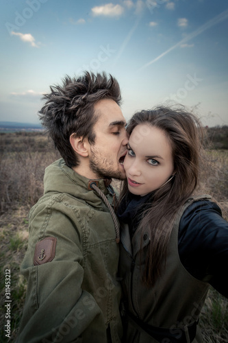 Portrait of young man, who is biting woman's face. Woman is looking in to camera. Couple is stretching hands out of picture. Background out of focus with withered vegetation and blue sky. © Zdena Venclik