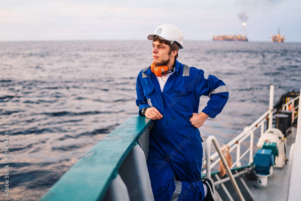 Naklejka premium Marine Deck Officer or Chief mate seaman on deck of offshore vessel or ship , wearing PPE personal protective equipment - helmet, coverall looks at sea. ocean view
