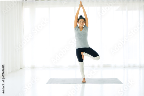 Asian woman practicing yoga at home, standing in Vrksasana or Tree pose hands above the head, working out, wearing sportswear, concept of healthy life and natural balance