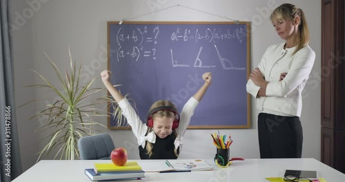 Little Caucasian girl sitting at the table in headphones and showing thumbs up as adult tutor standing next to her. Schoolgirl misbehaving studying at home. Education, learning. Cinema 4k ProRes HQ. photo