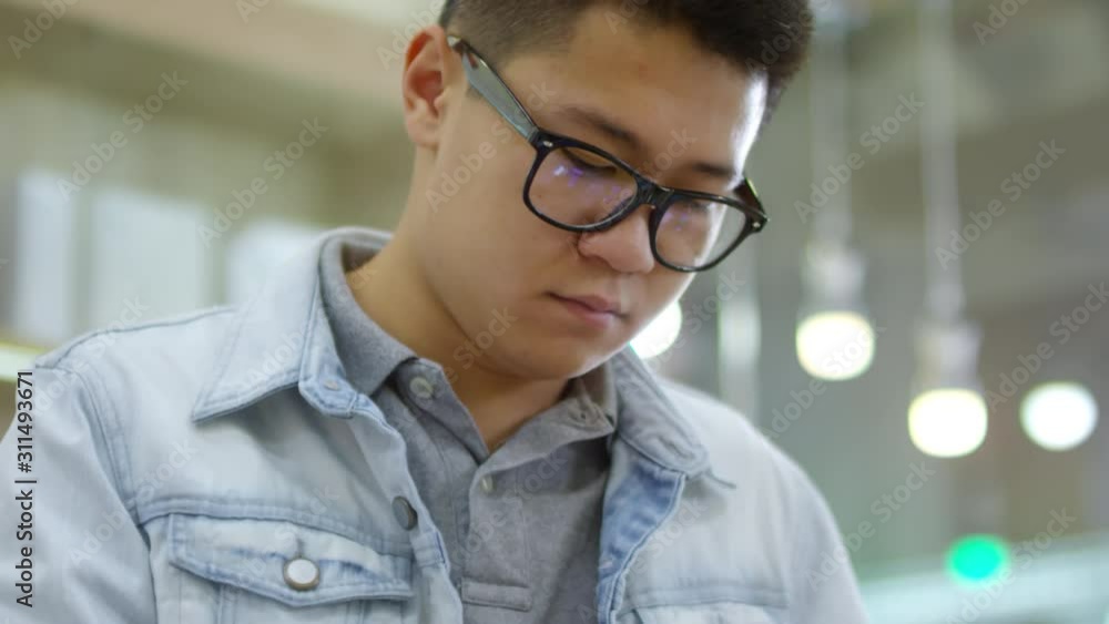 Tilt down shot of young Asian man in glasses choosing and testing smartphones at gadget store, handheld Tilt down shot of young Asian man in glasses choosing and testing smartphones at gadget store, handheld