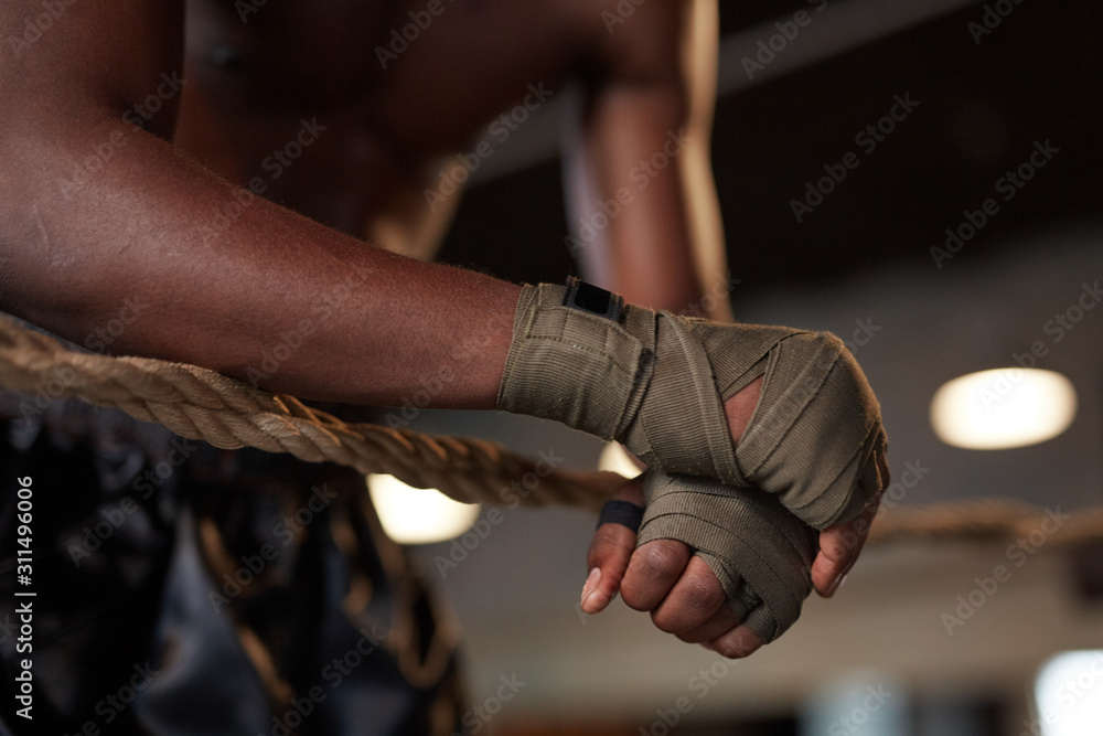 Fototapeta premium Close-up of African boxer in bandage on his hands leaning on the ropes of boxing ring in the gym