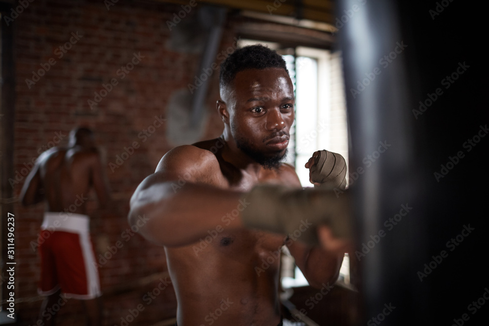 Fototapeta premium African serious boxer fighting with punching bag in gym he preparing for competition with his partner in the background