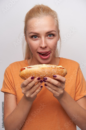 Close-up of hungry blue-eyed excited young blonde lady with tasty hot dog in her hands looking cheerfully at camera and licking her lips, standing against white background