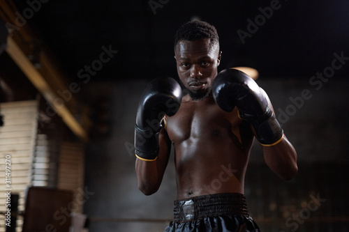 Portrait of African muscular boxer in boxing gloves standing in fight pose and looking at camera