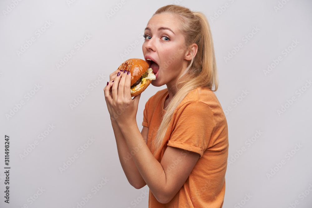 Studio photo of pretty young long haired blonde woman in casual wear holding tasty hamburger in her hands and going to eat it, standing against white background Studio photo of pretty young long haired blonde woman in casual wear holding tasty hamburger in her hands and going to eat it, standing against white background