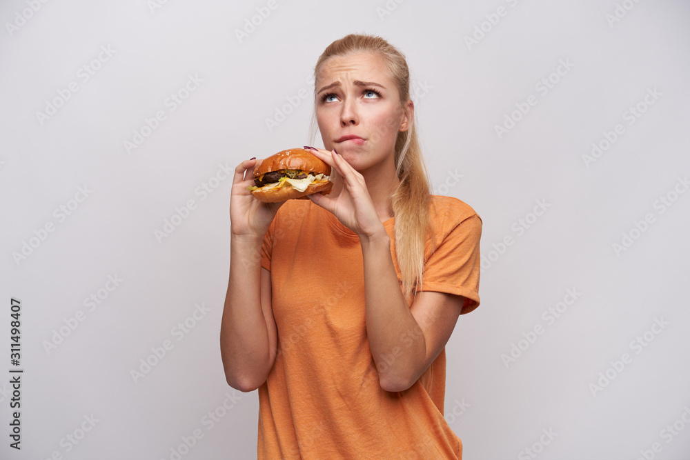 Indoor photo of young attractive blonde woman with ponytail hairstyle feeling guilty while eating unhealthy food, looking doubtful upwards and frowning her eyebrows over white background Indoor photo of young attractive blonde woman with ponytail hairstyle feeling guilty while eating unhealthy food, looking doubtful upwards and frowning her eyebrows over white background