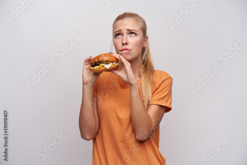 Indoor photo of young attractive blonde woman with ponytail hairstyle feeling guilty while eating unhealthy food  looking doubtful upwards and frowning her eyebrows over white background