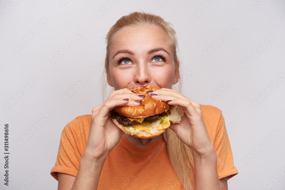 Portrait of pleased young lovely blonde woman with casual hairstyle eating fresh hamburger with great appetite and looking cheerfully upwards, posing over white background