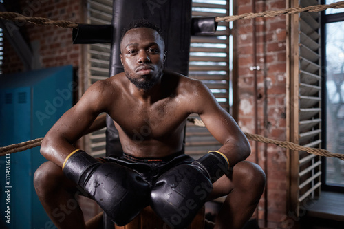 Portrait of African young shirtless boxer in boxing gloves looking at camera while sitting and resting on boxing ring