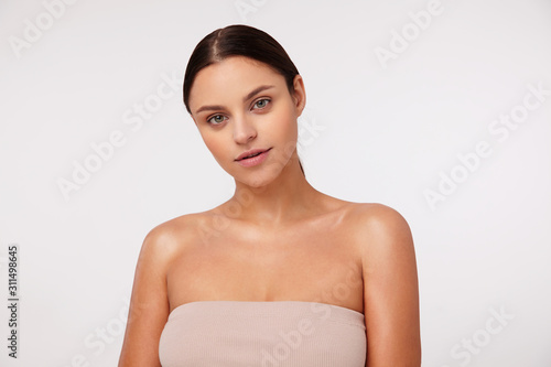 Portrait of beautiful young dark haired female with casual hairstyle looking positively at camera with calm face, posing over white background in beige top with opened shoulders