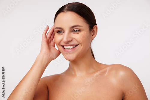 Cheerful lovely young brunette woman with ponytail hairstyle keeping raised hand behind her ear and smiling happily while posing against white background