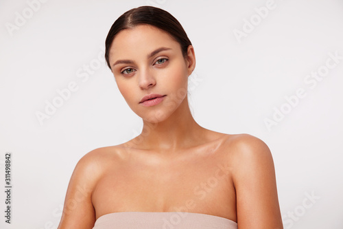 Pensive young attractive brunette female with natural makeup wearing casual hairstyle while standing against white background, dressed in nude top with opened shoulders