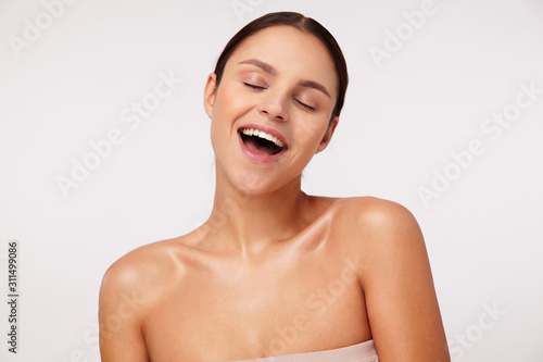 Indoor photo of pleasant looking pretty young brunette female wearing natural makeup while standing over white background, rejoicing about something positive with closed eyes