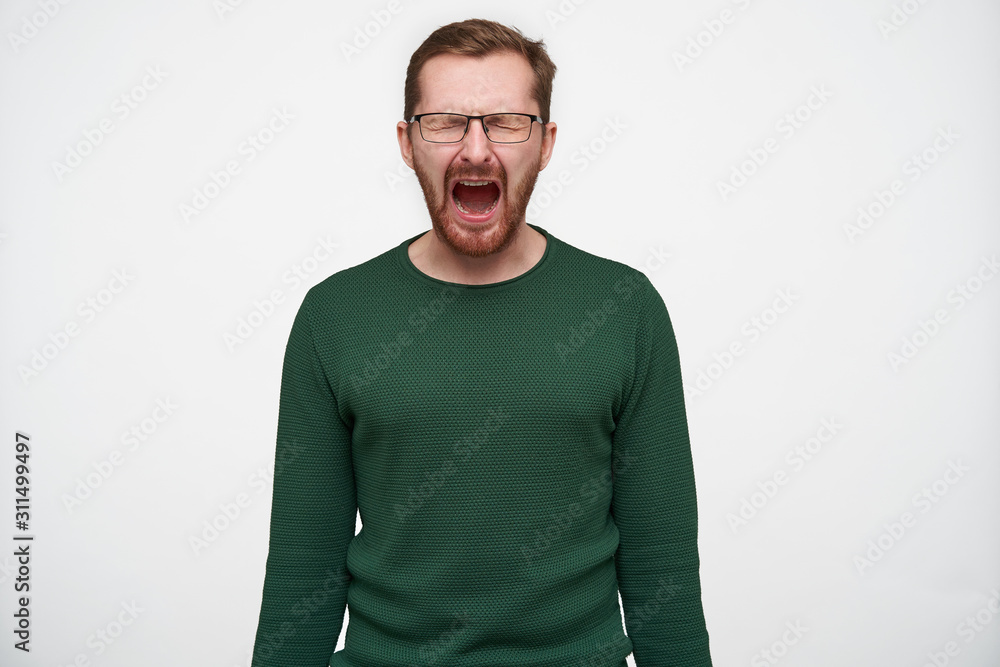 Stressed young short haired brunette man with beard wearing eyewear and green sweater posing over white background with hands down, screaming desperately with closed eyes