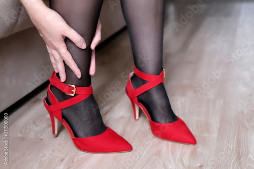 Tired legs, foot pain, woman in red shoes on high heels massaging her leg after work. Girl in black stockings, female health and fashion