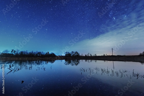 A magical starry night on the river bank with a large tree and a milky way in the sky and falling stars in the summer. 