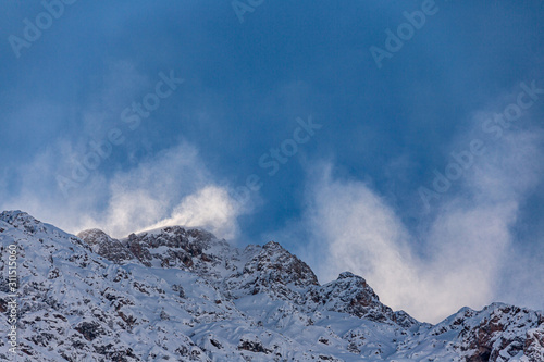 Snow dust in the sun in the air on the top of a mountain against a blue sky. Strong wind on top of the mountain. The wind tears the snow cover from the top of mount. Mountain peak in the cloud. © Relisa