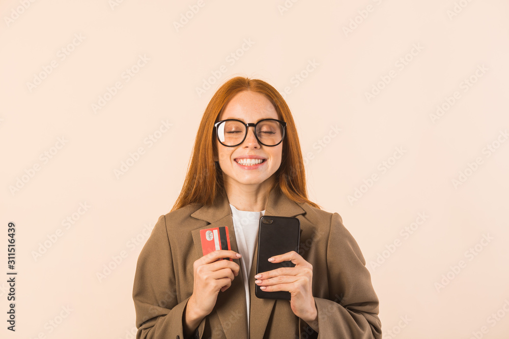 beautiful young woman with red hair in a jacket on a beige background with a credit card and a phone in her hands beautiful young woman with red hair in a jacket on a beige background with a credit card and a phone in her hands